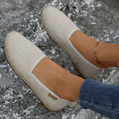 Close-up of a woman's hand holding a pair of slip-on shoes with a textured fabric design. One shoe is on her foot, showcasing a comfortable and casual style ideal for everyday wear.