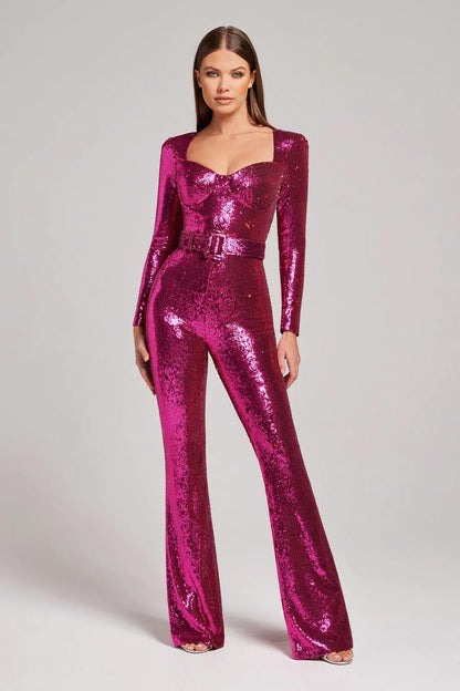 Woman wearing the Serena Women's Sequin Jumpsuit, a glamorous wide-leg jumpsuit with a shimmering sequin design, posing elegantly on a terrace at night with architectural buildings in the background.