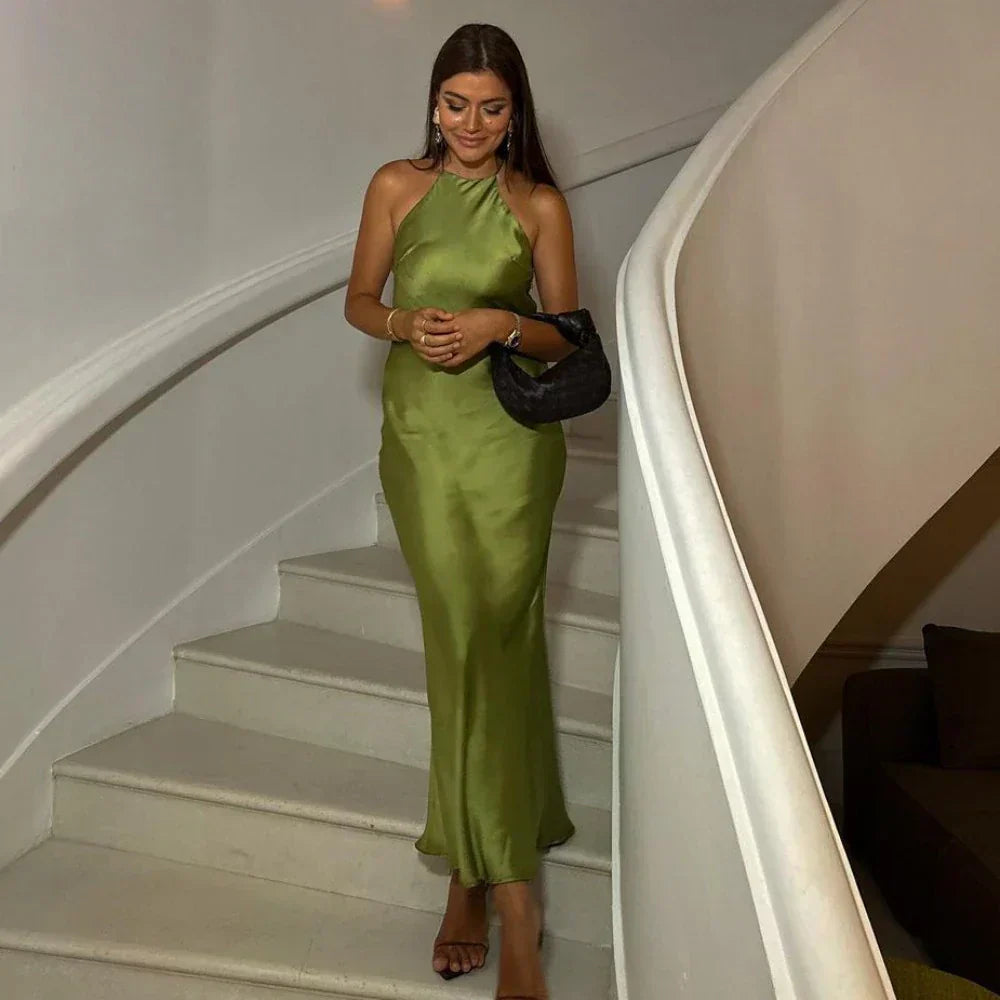 Elegant woman wearing a satin halter maxi dress, descending a staircase with a small handbag in hand, showcasing the dress's flowing silhouette and refined design.