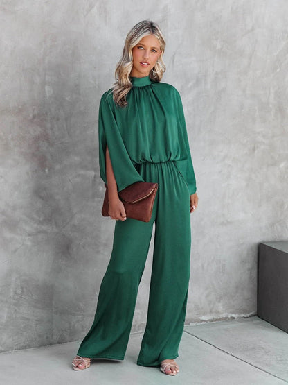 A woman wearing a high-neck, wide-leg dress with flowing sleeves, styled elegantly with a clutch bag. The dress features a relaxed silhouette that combines sophistication with comfort, perfect for both casual and formal occasions.