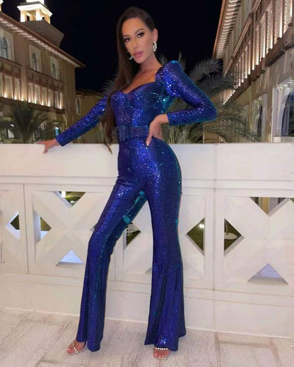 Woman wearing the Serena Women's Sequin Jumpsuit, a glamorous wide-leg jumpsuit with a shimmering sequin design, posing elegantly on a terrace at night with architectural buildings in the background.