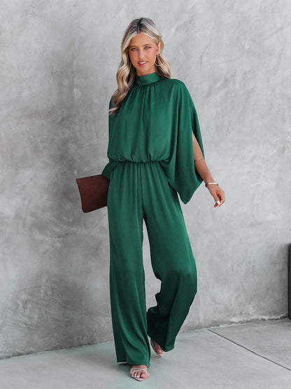 A woman wearing a high-neck, wide-leg dress with flowing sleeves, styled elegantly with a clutch bag. The dress features a relaxed silhouette that combines sophistication with comfort, perfect for both casual and formal occasions.