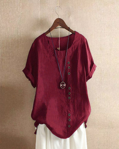 A casual women's blouse with an asymmetrical button design, short sleeves, and a relaxed fit, hanging on a wooden hanger. This blouse, part of the Mira collection, offers a unique style perfect for casual and everyday wear.