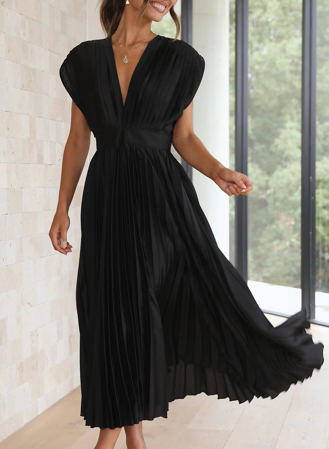 A woman wearing a pleated midi dress with a deep V-neck design, paired with a clutch and subtle jewelry, standing indoors. The dress features a fitted waist and flowing pleats, creating an elegant and refined silhouette perfect for formal events or special occasions.