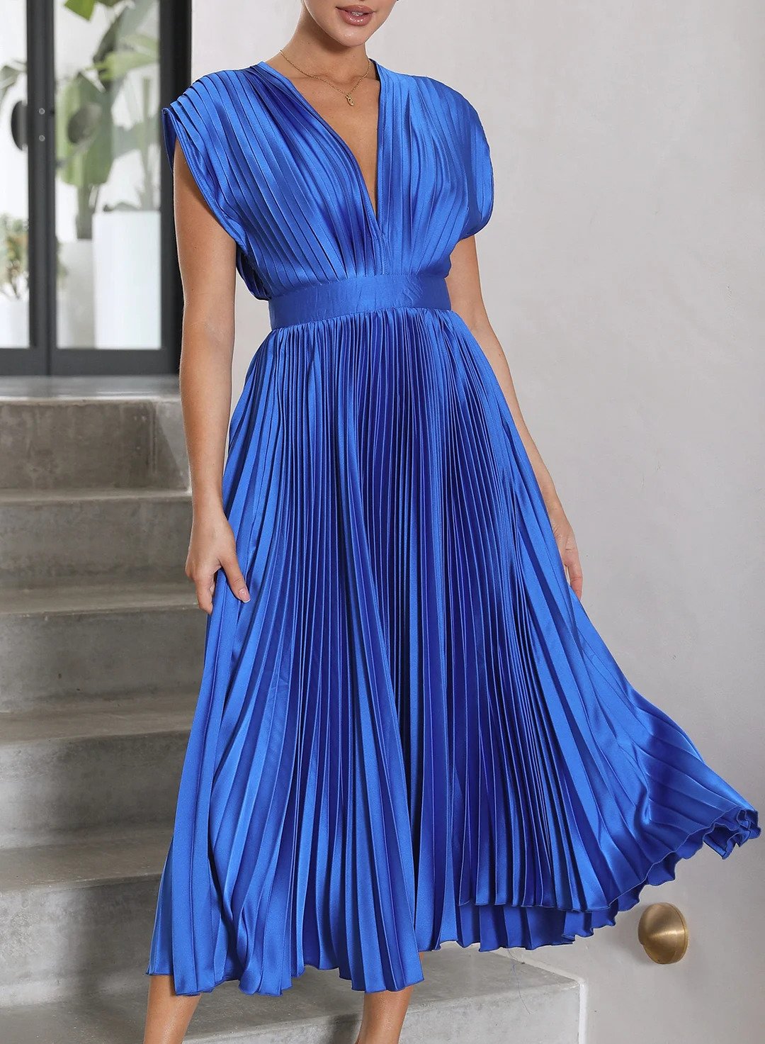 A woman wearing a pleated midi dress with a deep V-neck design, paired with a clutch and subtle jewelry, standing indoors. The dress features a fitted waist and flowing pleats, creating an elegant and refined silhouette perfect for formal events or special occasions.