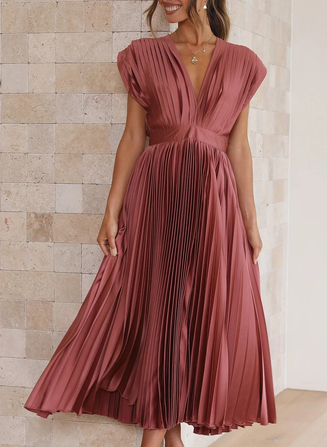 A woman wearing a pleated midi dress with a deep V-neck design, paired with a clutch and subtle jewelry, standing indoors. The dress features a fitted waist and flowing pleats, creating an elegant and refined silhouette perfect for formal events or special occasions.