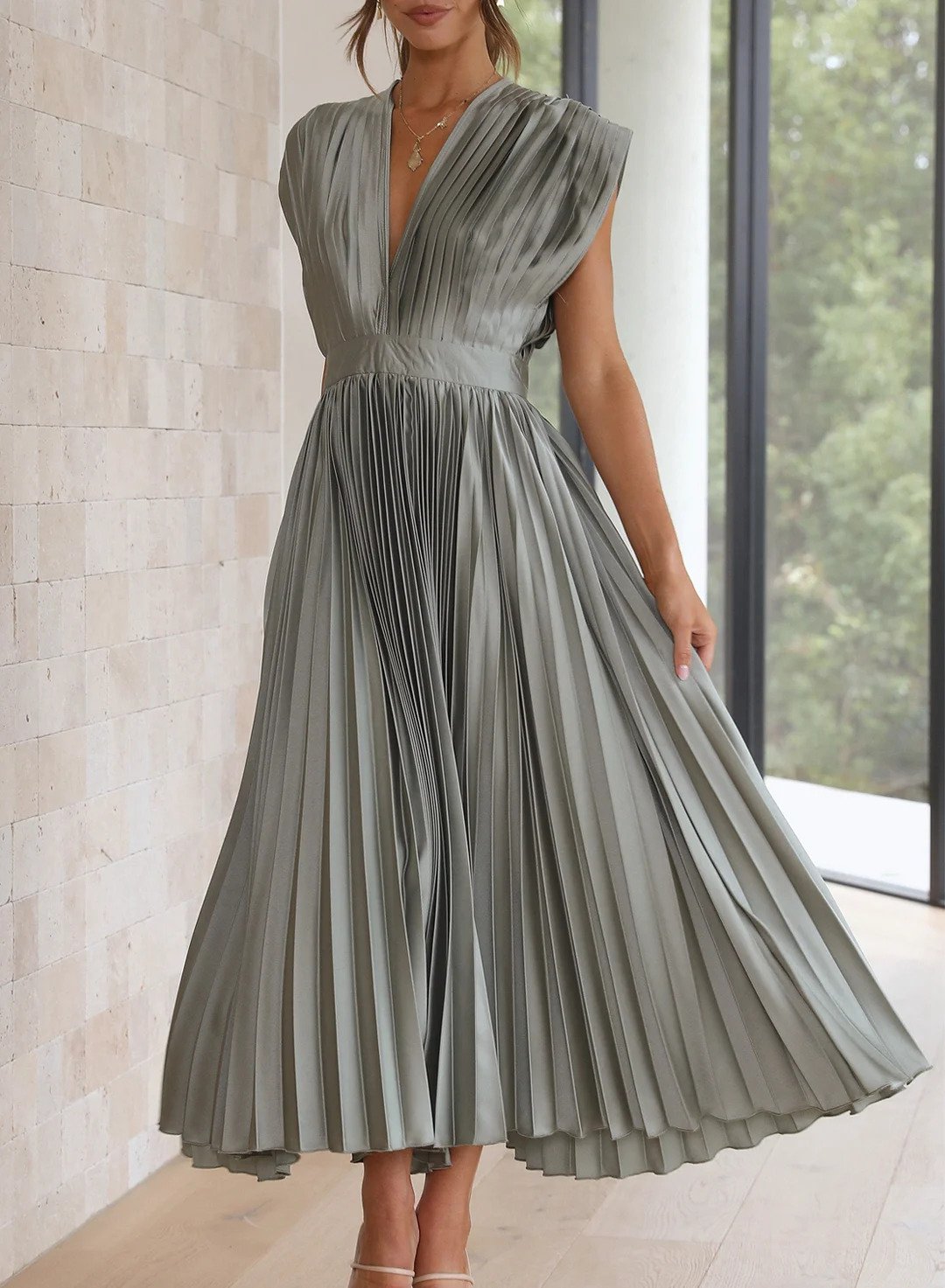 A woman wearing a pleated midi dress with a deep V-neck design, paired with a clutch and subtle jewelry, standing indoors. The dress features a fitted waist and flowing pleats, creating an elegant and refined silhouette perfect for formal events or special occasions.