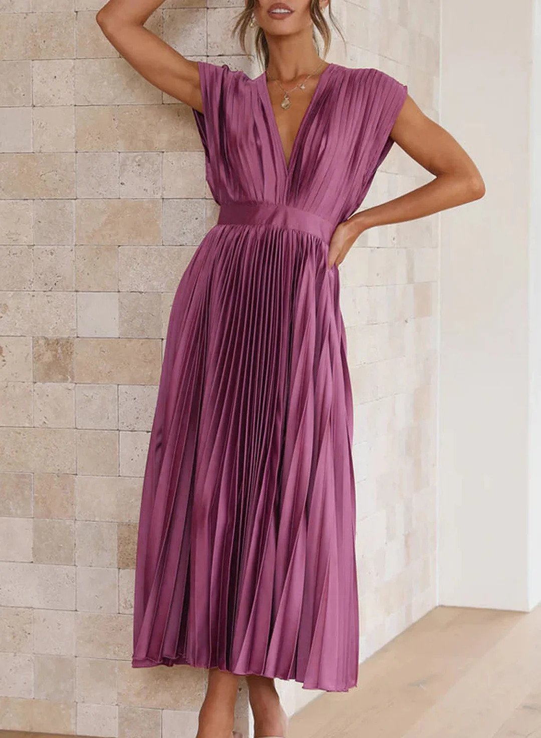 A woman wearing a pleated midi dress with a deep V-neck design, paired with a clutch and subtle jewelry, standing indoors. The dress features a fitted waist and flowing pleats, creating an elegant and refined silhouette perfect for formal events or special occasions.