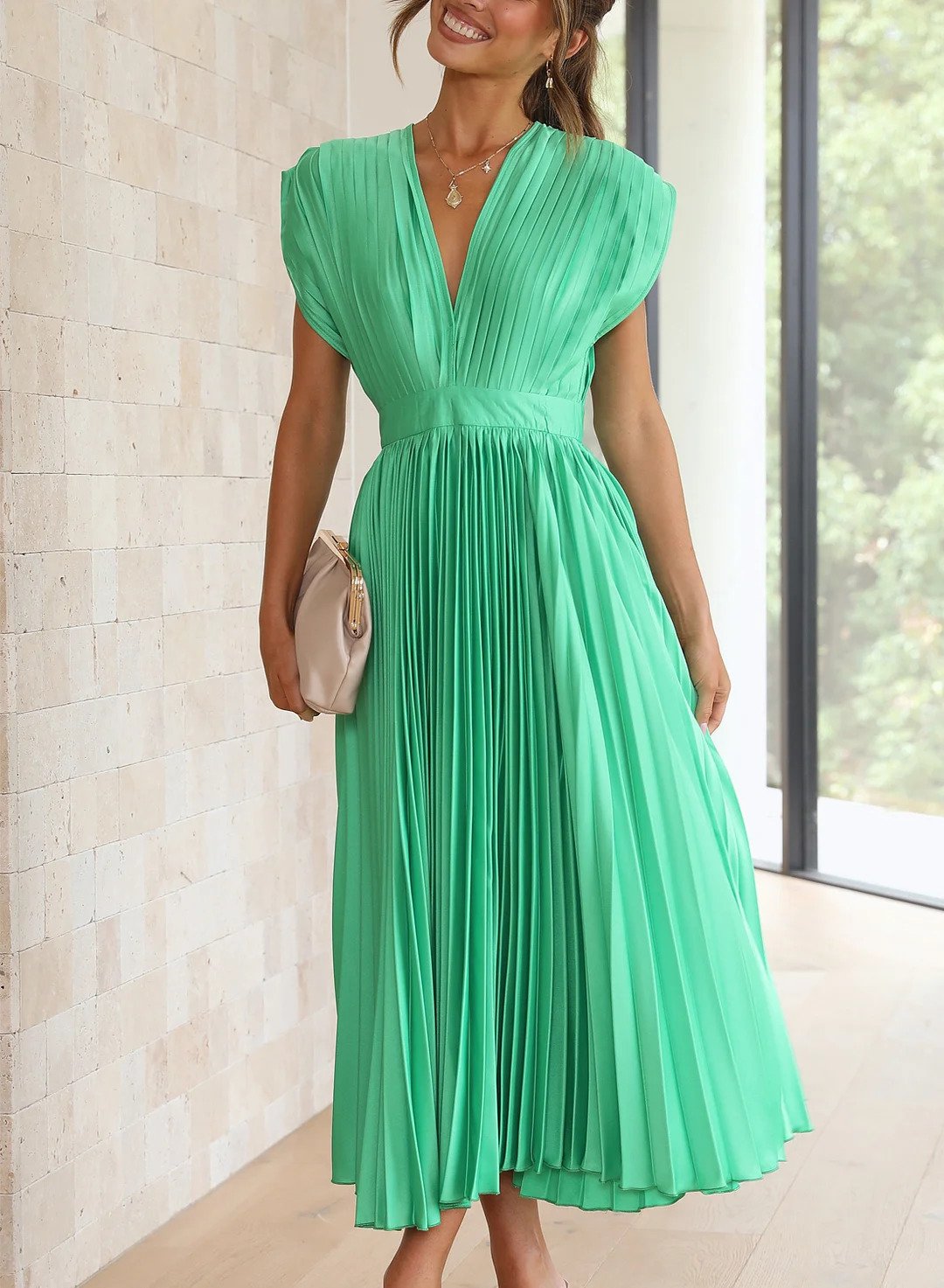 A woman wearing a pleated midi dress with a deep V-neck design, paired with a clutch and subtle jewelry, standing indoors. The dress features a fitted waist and flowing pleats, creating an elegant and refined silhouette perfect for formal events or special occasions.