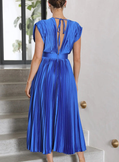 A woman wearing a pleated midi dress with a deep V-neck design, paired with a clutch and subtle jewelry, standing indoors. The dress features a fitted waist and flowing pleats, creating an elegant and refined silhouette perfect for formal events or special occasions.