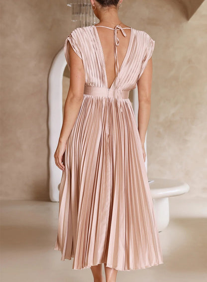 A woman wearing a pleated midi dress with a deep V-neck design, paired with a clutch and subtle jewelry, standing indoors. The dress features a fitted waist and flowing pleats, creating an elegant and refined silhouette perfect for formal events or special occasions.