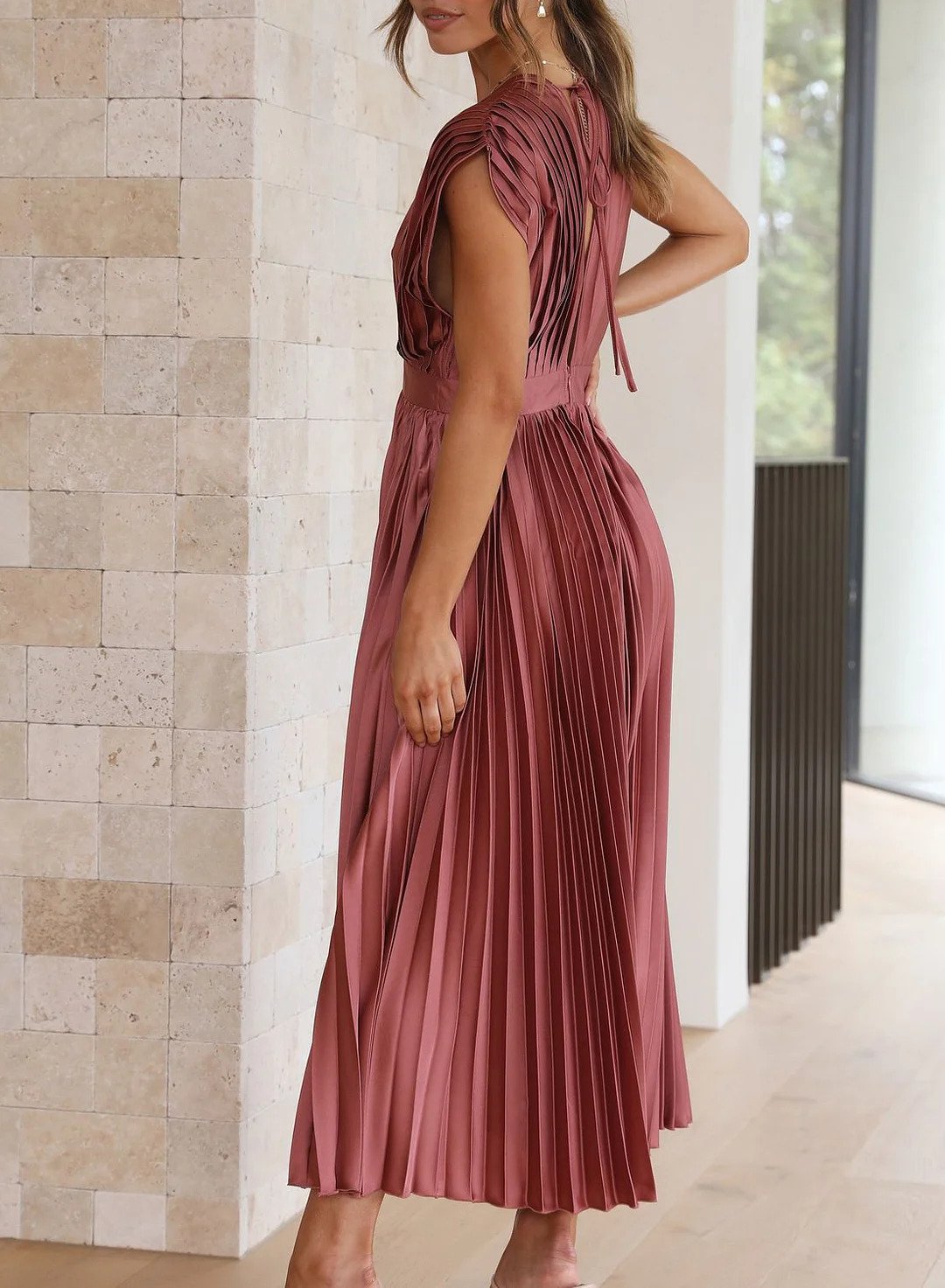 A woman wearing a pleated midi dress with a deep V-neck design, paired with a clutch and subtle jewelry, standing indoors. The dress features a fitted waist and flowing pleats, creating an elegant and refined silhouette perfect for formal events or special occasions.