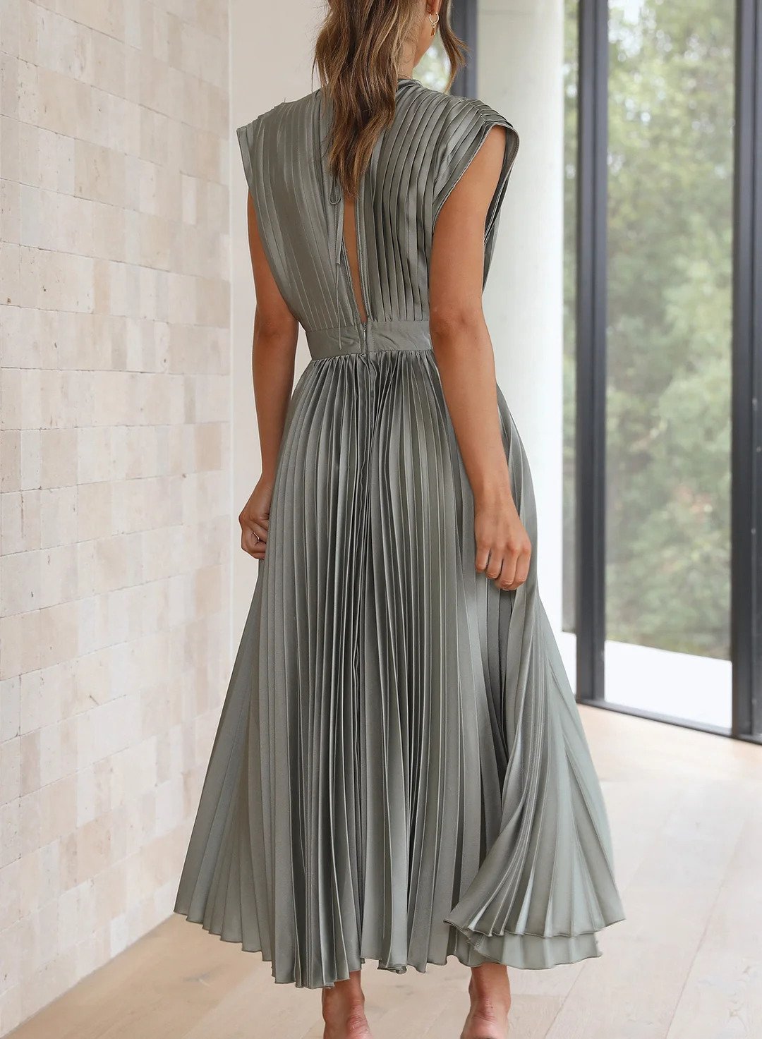 A woman wearing a pleated midi dress with a deep V-neck design, paired with a clutch and subtle jewelry, standing indoors. The dress features a fitted waist and flowing pleats, creating an elegant and refined silhouette perfect for formal events or special occasions.