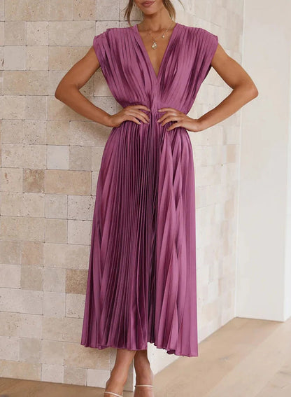 A woman wearing a pleated midi dress with a deep V-neck design, paired with a clutch and subtle jewelry, standing indoors. The dress features a fitted waist and flowing pleats, creating an elegant and refined silhouette perfect for formal events or special occasions.