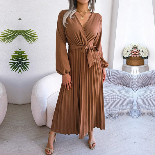 Woman wearing the Claire Women's Pleated Midi Dress, a long-sleeved dress with a pleated skirt and belted waist, gracefully holding the skirt to showcase its flow and elegance, standing in a stylish indoor setting.