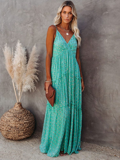 Woman wearing the Sienna Women's Tiered Maxi Dress, a long, flowy dress with adjustable straps and a bohemian-inspired tiered design, accessorized with layered necklaces and a clutch, posing gracefully against a textured wall with pampas grass in the background.
