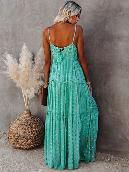Woman wearing the Sienna Women's Tiered Maxi Dress, a long, flowy dress with adjustable straps and a bohemian-inspired tiered design, accessorized with layered necklaces and a clutch, posing gracefully against a textured wall with pampas grass in the background.