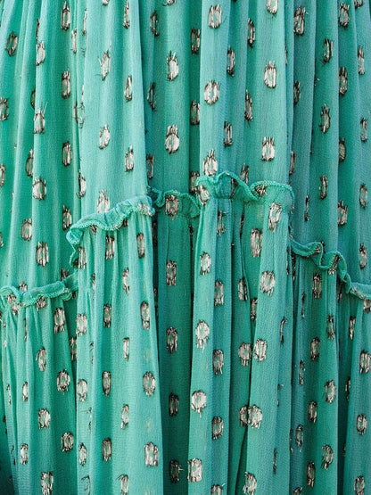 Woman wearing the Sienna Women's Tiered Maxi Dress, a long, flowy dress with adjustable straps and a bohemian-inspired tiered design, accessorized with layered necklaces and a clutch, posing gracefully against a textured wall with pampas grass in the background.