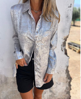 Woman wearing the Sophia Women's Shimmering Button-Up Blouse, a relaxed-fit blouse with a soft shimmer and long sleeves, styled with casual shorts and minimal jewelry, standing against a textured wall.