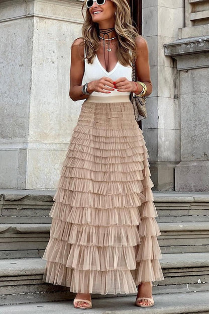 A woman sitting on outdoor steps, wearing a flowing ruffled midi skirt with layered tulle details. She pairs it with a sleeveless top, heels, and a chic handbag, creating an elegant and feminine look ideal for casual outings or dressy occasions.