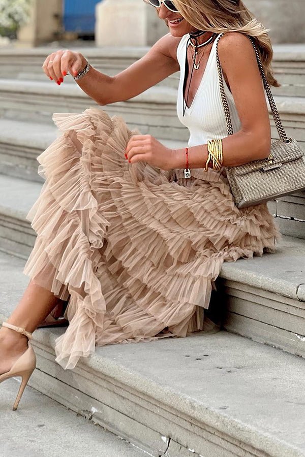 A woman sitting on outdoor steps, wearing a flowing ruffled midi skirt with layered tulle details. She pairs it with a sleeveless top, heels, and a chic handbag, creating an elegant and feminine look ideal for casual outings or dressy occasions.