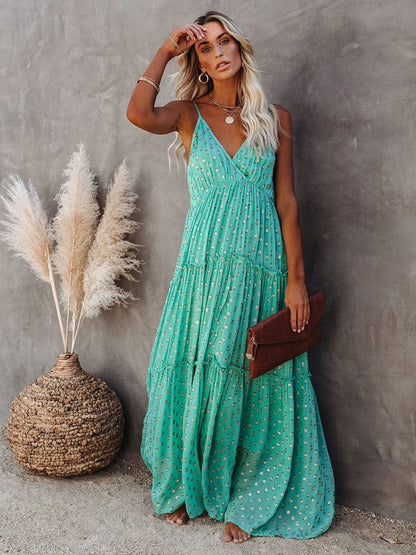 Woman wearing the Sienna Women's Tiered Maxi Dress, a long, flowy dress with adjustable straps and a bohemian-inspired tiered design, accessorized with layered necklaces and a clutch, posing gracefully against a textured wall with pampas grass in the background.