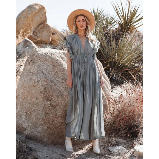 A woman wearing the Amara Women's Flowing Maxi Dress, styled with a wide-brimmed hat and ankle boots, standing outdoors among desert rocks. The dress has a boho-chic design with a relaxed, flowing silhouette, perfect for a free-spirited, elegant look.