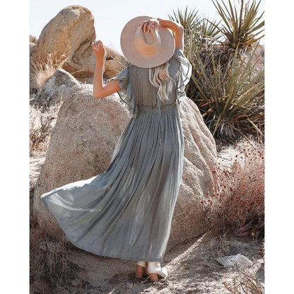 A woman wearing the Amara Women's Flowing Maxi Dress, styled with a wide-brimmed hat and ankle boots, standing outdoors among desert rocks. The dress has a boho-chic design with a relaxed, flowing silhouette, perfect for a free-spirited, elegant look.