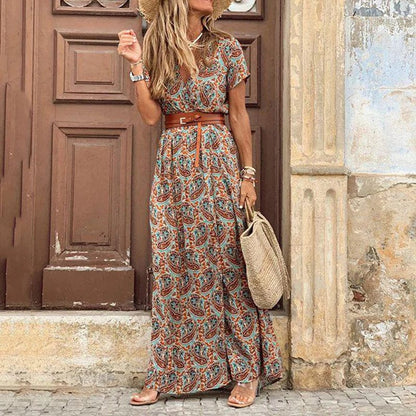 A woman wearing the Layla | Bohemian Print Maxi Dress, accessorized with a stylish belt and woven handbag, standing against a rustic door backdrop. The dress’s flowy and bohemian-inspired design makes it perfect for casual outings or a relaxed, chic look for the day.