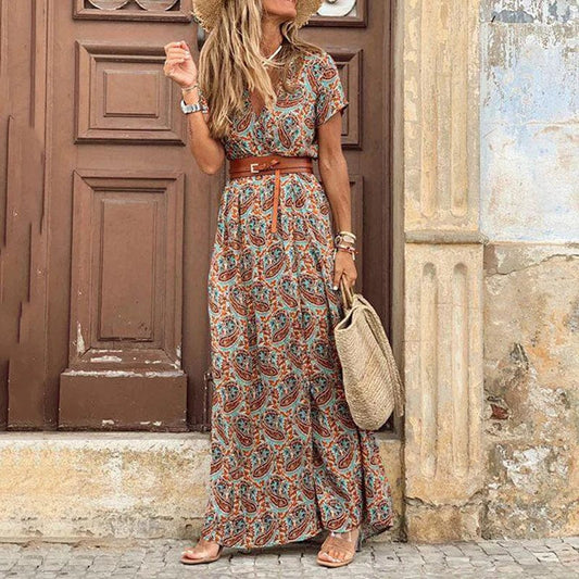 A woman wearing the Layla | Bohemian Print Maxi Dress, accessorized with a stylish belt and woven handbag, standing against a rustic door backdrop. The dress’s flowy and bohemian-inspired design makes it perfect for casual outings or a relaxed, chic look for the day.