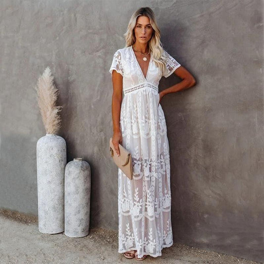 Woman wearing the Aurora Women's Lace Maxi Dress, a long dress with intricate lace detailing and a sheer, elegant design, standing against a textured wall with decorative vases nearby, exuding a graceful and sophisticated look.