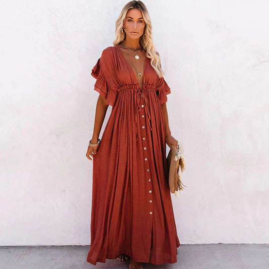 A woman sitting in a woven chair wearing a flowing boho maxi dress with a button-down front and cinched waist. The dress features a relaxed, loose fit and short sleeves, creating an effortlessly stylish and laid-back look. She accessorizes with layered necklaces and a chic handbag.