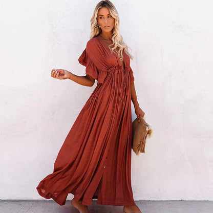 A woman sitting in a woven chair wearing a flowing boho maxi dress with a button-down front and cinched waist. The dress features a relaxed, loose fit and short sleeves, creating an effortlessly stylish and laid-back look. She accessorizes with layered necklaces and a chic handbag.