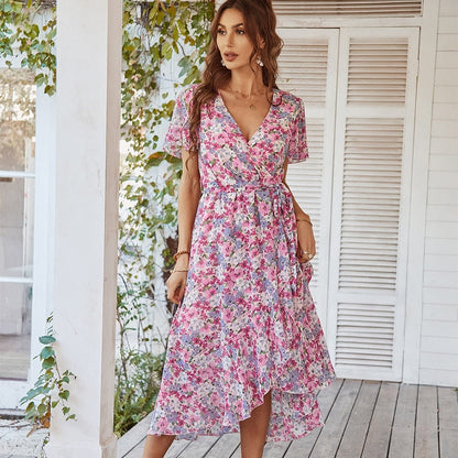 A woman wearing the Clara Women's Elegant Wrap Dress, a floral midi dress with a flattering wrap design, short sleeves, and an asymmetrical hemline, standing on a wooden porch surrounded by greenery.