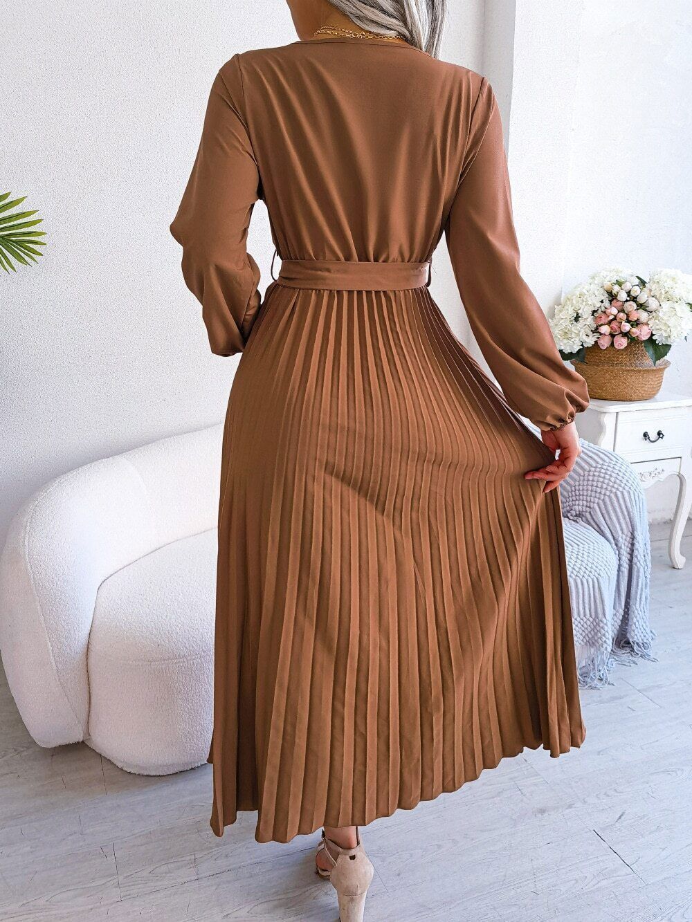 Woman wearing the Claire Women's Pleated Midi Dress, a long-sleeved dress with a pleated skirt and belted waist, gracefully holding the skirt to showcase its flow and elegance, standing in a stylish indoor setting.