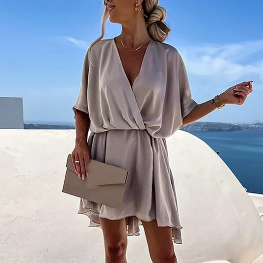 A woman wearing a flowy wrap dress with a V-neck design, posing outdoors against a scenic background. The dress features a relaxed fit with short sleeves, perfect for both casual and elegant occasions. She holds a stylish clutch, complementing the effortlessly chic look.