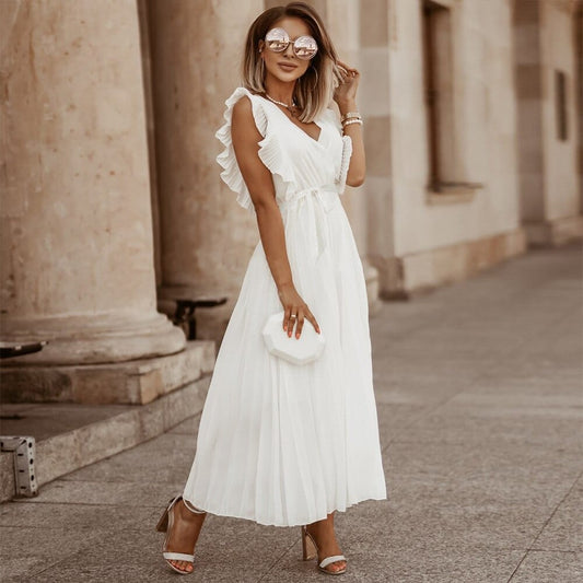 A woman wearing a stylish maxi dress with ruffle details on the shoulders and a pleated skirt, accessorized with a small clutch and heels, posing outdoors in front of elegant architecture. The dress has a flattering, flowy silhouette perfect for formal events or casual outings.